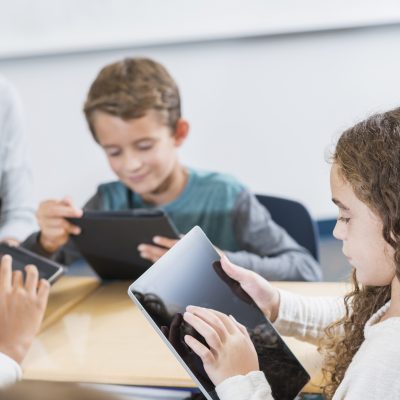Co2 controle in klaslokalen is goed voor de leerprestaties Een sfeerbeeld van een groep kinderen die leren met een tablet en de lerares lachtend mee kijkt.